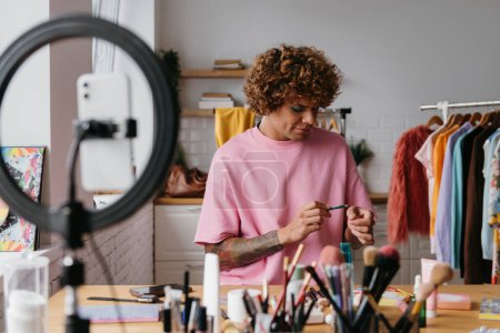 Téléchargez les photos : Confiant jeune homme gay examiner des produits de beauté tout en enregistrant tutoriel maquillage de la maison - en image libre de droit