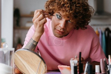 Foto de Confiado joven gay hombre aplicación de maquillaje y mirando a la cámara, mientras que pasar tiempo en casa - Imagen libre de derechos