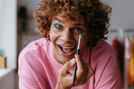 Photo for Happy young gay man holding make-up brush while applying eyeshadow at home - Royalty Free Image