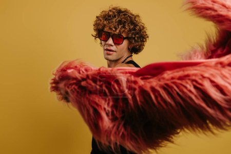 Photo for Curly young gay man in trendy glasses playing with fluffy coat against yellow background - Royalty Free Image