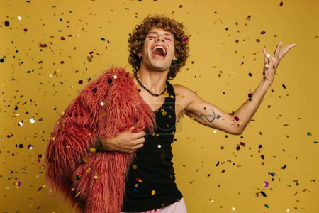 Téléchargez les photos : Joyeux jeune homme gay portant un manteau moelleux et jetant des confettis colorés sur fond jaune - en image libre de droit