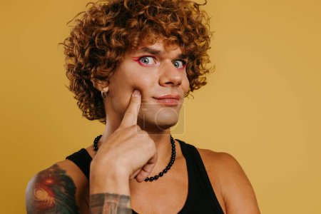 Téléchargez les photos : Portrait de jeune homme gay ludique avec maquillage touchant visage sur fond jaune - en image libre de droit