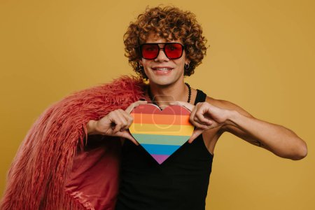 Téléchargez les photos : Jeune homme gay à la mode tenant le drapeau arc-en-ciel en forme de coeur et souriant sur fond jaune - en image libre de droit