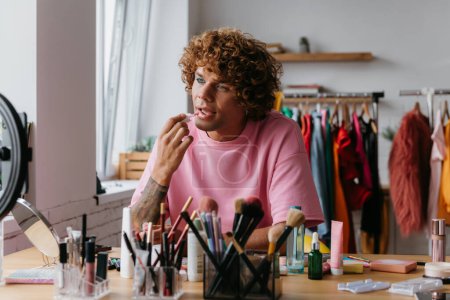 Photo for Confident young gay man applying lipstick while streaming make-up vlog from home - Royalty Free Image
