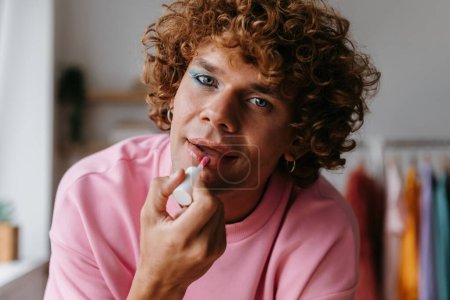 Photo for Young gay man with make-up applying lipstick and looking at camera while spending time at home - Royalty Free Image