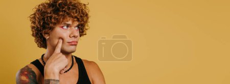 Téléchargez les photos : Portrait de jeune homme gay avec maquillage toucher le visage sur fond jaune - en image libre de droit