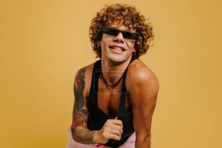 Téléchargez les photos : Joyeux jeune homme gay en débardeur et des lunettes à la mode souriant sur fond jaune - en image libre de droit