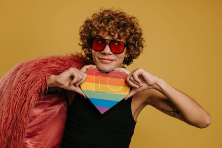 Téléchargez les photos : Jeune homme gay ludique tenant le drapeau arc-en-ciel en forme de coeur et souriant sur fond jaune - en image libre de droit