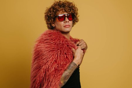 Téléchargez les photos : Bouclé jeune gay homme dans des lunettes à la mode portant manteau moelleux sur fond jaune - en image libre de droit