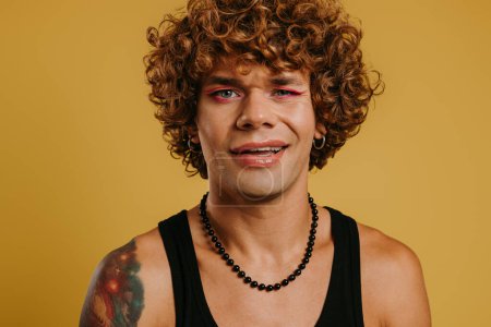 Téléchargez les photos : Portrait de beau jeune homme gay avec maquillage clin d'oeil à la caméra sur fond jaune - en image libre de droit
