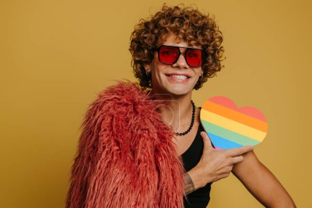 Foto de Feliz joven gay hombre usando mullido abrigo y sosteniendo corazón en forma de arco iris bandera en amarillo fondo - Imagen libre de derechos