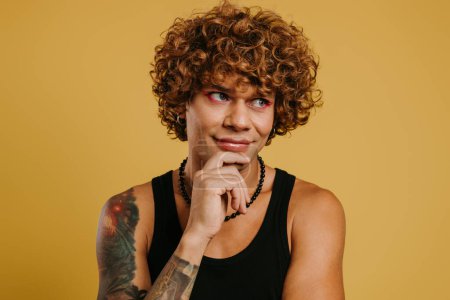 Téléchargez les photos : Portrait de jeune homme gay mignon avec maquillage tenant la main sur le menton sur fond jaune - en image libre de droit