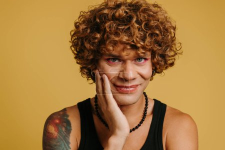 Téléchargez les photos : Portrait de beau jeune homme gay avec maquillage touchant visage sur fond jaune - en image libre de droit