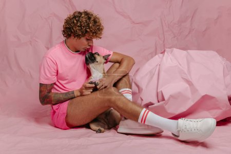 Téléchargez les photos : Élégant jeune gay l'homme caresser son mignon chien tout en étant assis contre froissé rose papier fond - en image libre de droit