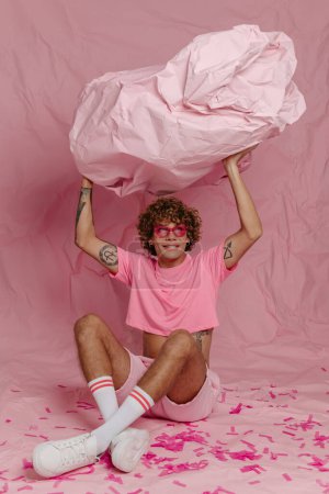 Téléchargez les photos : Élégant jeune gay l'homme dans à la mode porter sourire tout en étant couché sur froissé rose fond - en image libre de droit