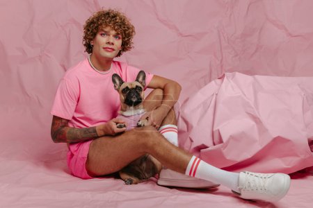 Photo for Stylish young gay man holding his cute dog while sitting against crumpled pink paper background - Royalty Free Image