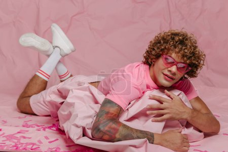 Téléchargez les photos : Élégant jeune gay l'homme dans à la mode usure penché sur wad de froissé rose papier tout en étant couché sur coloré fond - en image libre de droit