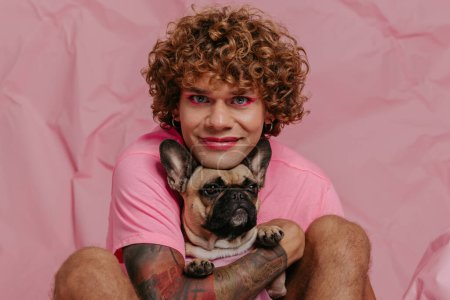 Téléchargez les photos : Beau jeune homme gay tenant son chien mignon et souriant tout en étant assis sur un fond rose froissé - en image libre de droit