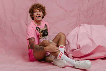 Foto de Moda joven gay hombre sosteniendo su lindo perro mientras sentado contra arrugado rosa fondo - Imagen libre de derechos