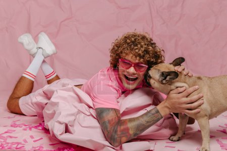 Téléchargez les photos : Élégant gay l'homme penché sur wad de froissé rose papier et caresser chien tout en étant couché sur coloré fond - en image libre de droit