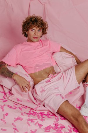 Foto de Vista superior de hombre gay joven con estilo en ropa rosa de moda mirando a la cámara mientras está sentado en papel de color arrugado - Imagen libre de derechos