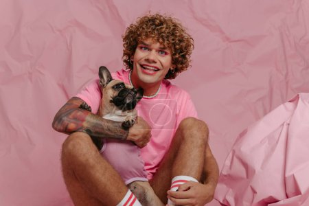 Foto de Elegante joven gay hombre sosteniendo su lindo perro y sonriendo mientras se sienta contra arrugado rosa fondo - Imagen libre de derechos