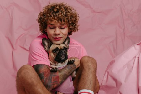 Téléchargez les photos : Élégant jeune homme gay embrassant son chien mignon et souriant tout en étant assis sur un fond rose froissé - en image libre de droit