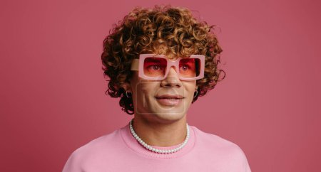 Téléchargez les photos : Jeune homme à la mode aux cheveux bouclés aux lunettes de style rétro souriant sur fond rose - en image libre de droit