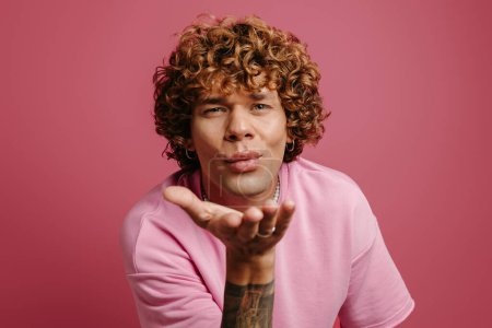 Téléchargez les photos : Beau jeune bouclé cheveux homme souffler un baiser sur caméra sur fond rose - en image libre de droit