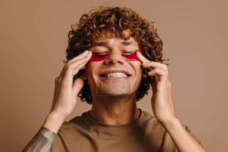 Téléchargez les photos : Homme heureux bénéficiant d'un traitement de beauté pour la peau tout en appliquant un masque cosmétique facial sur fond brun - en image libre de droit
