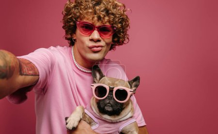 Téléchargez les photos : Cool jeune homme tenant français Bulldog animal de compagnie dans des lunettes funky et faire du selfie sur fond rose - en image libre de droit