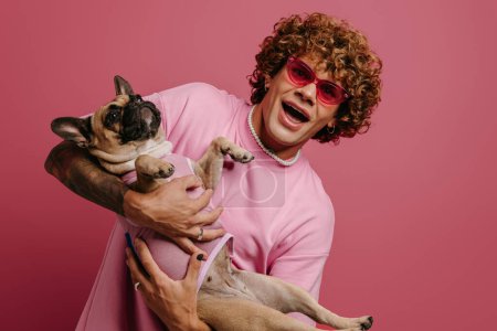 Téléchargez les photos : Jeune homme à la mode aux cheveux bouclés s'amusant avec son chien mignon et souriant sur fond rose - en image libre de droit