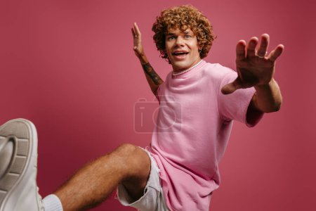 Photo for Joyful young curly hair man looking playful while kicking against pink background - Royalty Free Image