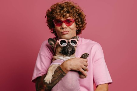 Foto de Juguetón joven pelo rizado hombre celebración Bulldog francés mascota en gafas funky y muecas sobre fondo rosa - Imagen libre de derechos