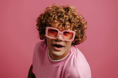 Téléchargez les photos : Mode jeune bouclé cheveux gay homme dans rétro lunettes de vue de style regardant caméra sur fond rose - en image libre de droit