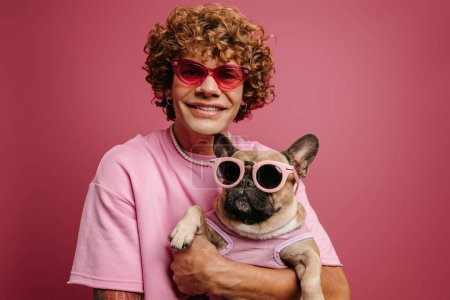 Photo for Fashionable young curly hair man holding French Bulldog pet in funky glasses on pink background - Royalty Free Image