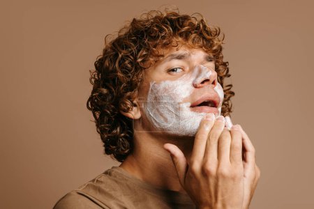 Téléchargez les photos : Beau jeune homme bénéficiant d'un traitement de beauté tout en appliquant de la crème sur le visage et debout sur fond brun - en image libre de droit