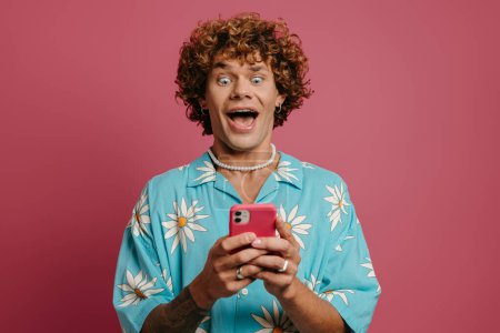 Téléchargez les photos : Surpris jeune homme aux cheveux bouclés en chemise funky regardant son téléphone intelligent sur fond rose - en image libre de droit