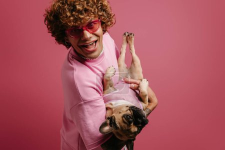 Foto de Moda joven pelo rizado hombre divirtiéndose con su lindo perro y sonriendo sobre fondo rosa - Imagen libre de derechos