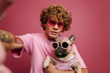 Téléchargez les photos : Jeune homme à la mode tenant un bouledogue français dans des lunettes funky et faisant du selfie sur fond rose - en image libre de droit