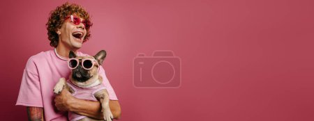 Téléchargez les photos : Joyeux jeune homme aux cheveux bouclés tenant un bouledogue français dans des lunettes funky sur fond rose - en image libre de droit