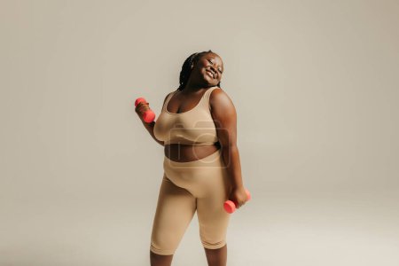 Téléchargez les photos : Confiant plus la taille de la femme africaine en vêtements de sport exercice avec haltères sur fond de studio - en image libre de droit