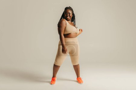 Foto de Feliz mujer africana con curvas en ropa deportiva que mira con confianza en su cuerpo mientras está de pie en el fondo del estudio - Imagen libre de derechos