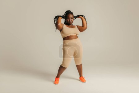Téléchargez les photos : Pleine longueur de belle africaine plus taille femme en vêtements de sport ajuster ses dreadlocs sur fond de studio - en image libre de droit