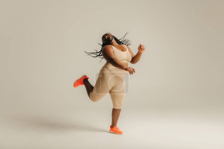 Téléchargez les photos : Toute la longueur de femme africaine courbée heureuse en tenue de sport dansant sur fond de studio - en image libre de droit