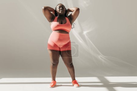 Foto de Longitud completa de mujer africana de tamaño grande en ropa deportiva ajustando el cabello mientras está de pie en el estudio de fitness - Imagen libre de derechos
