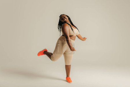 Téléchargez les photos : Pleine longueur heureux plus la taille femme africaine en vêtements de sport danse sur fond de studio - en image libre de droit