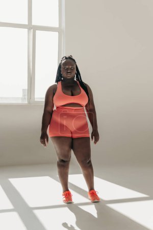 Téléchargez les photos : Pleine longueur de confiance africaine plus la taille femme en tenue de sport debout dans le studio de remise en forme - en image libre de droit