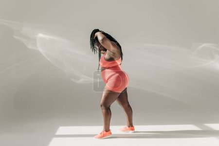 Téléchargez les photos : Pleine longueur de femme africaine plus la taille en vêtements de sport ajuster ses dreadlocs avant l'entraînement dans un studio de fitness - en image libre de droit