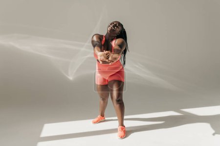 Téléchargez les photos : Pleine longueur de joyeux Africaine plus taille femme en vêtements de sport faire des exercices d'étirement dans un studio de fitness - en image libre de droit
